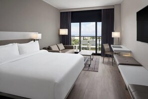 A hotel room with a large bed, a desk, a chair, and a view of the city. - AC Hotel Kingston Jamaica (Kingston)