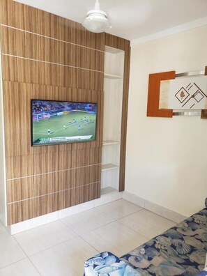 A living room with a flat-screen TV mounted on a wooden panel wall, a ceiling fan, and a patterned sofa. - Flat no Lacqua Diroma seu Resort (Caldas Novas)