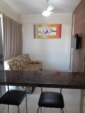 A modern kitchen with a granite countertop, two black bar stools, a ceiling fan, and a wall-mounted TV. - Flat no Lacqua Diroma seu Resort (Caldas Novas)