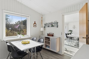 A dining area with a white table, black chairs, a window with a view of trees, and a wooden door leading to another room. - Spacious holiday home 150 m from the beach (Ålbæk)