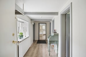 A hallway with a radiator, a door with a brass handle, a small table with a lamp, and a view into a room with a window and a chair.