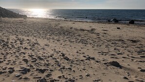Ranta, jossa näkyy selvästi jalanjälkiä ja jossa on kiviä, kirkas taivas ja aurinko, joka heijastuu vedenpinnasta.