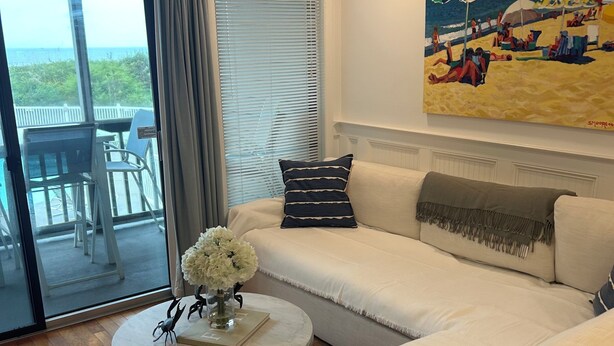En stue med en hvid sofa, et rundt sofabord med en vase blomster, et maleri af en strandscene og havudsigt gennem en skydedør.