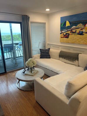 Uma sala de estar com sofá branco, mesa de centro redonda com um vaso de flores, um quadro de uma praia e vista para o oceano através de uma porta de correr de vidro.