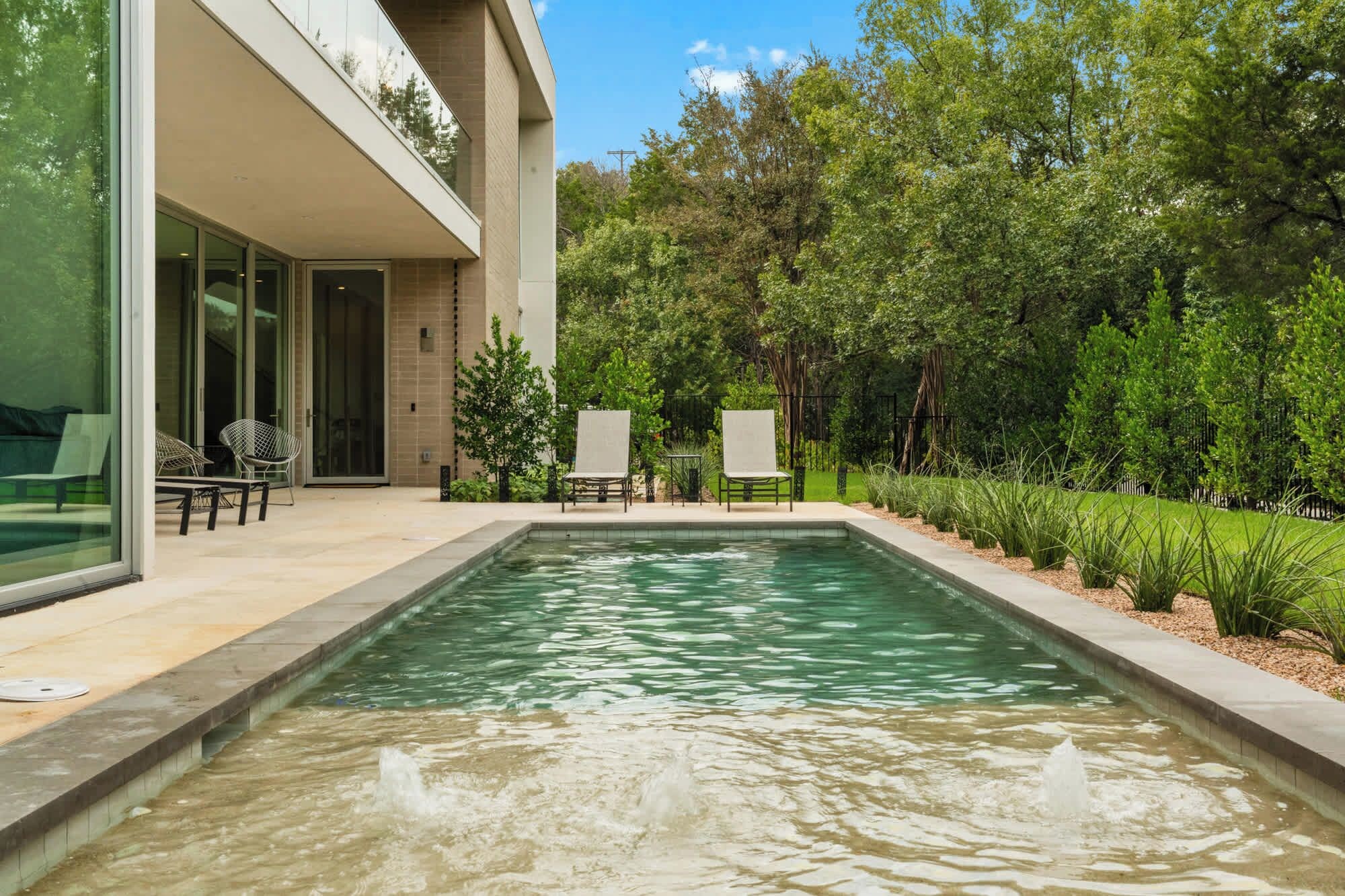 A swimming pool with a water feature, surrounded by a patio with chairs and a glass door leading to a covered area.