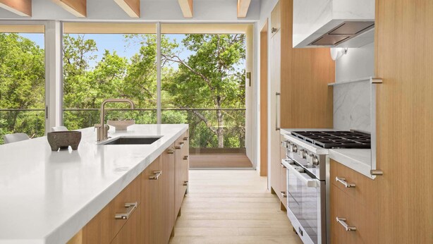 A modern kitchen with a large island, stainless steel appliances, and a view of trees through large windows.