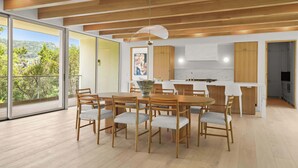 A modern kitchen with wooden cabinets, a dining area with a round table and chairs, and large windows offering a view of the outdoors.