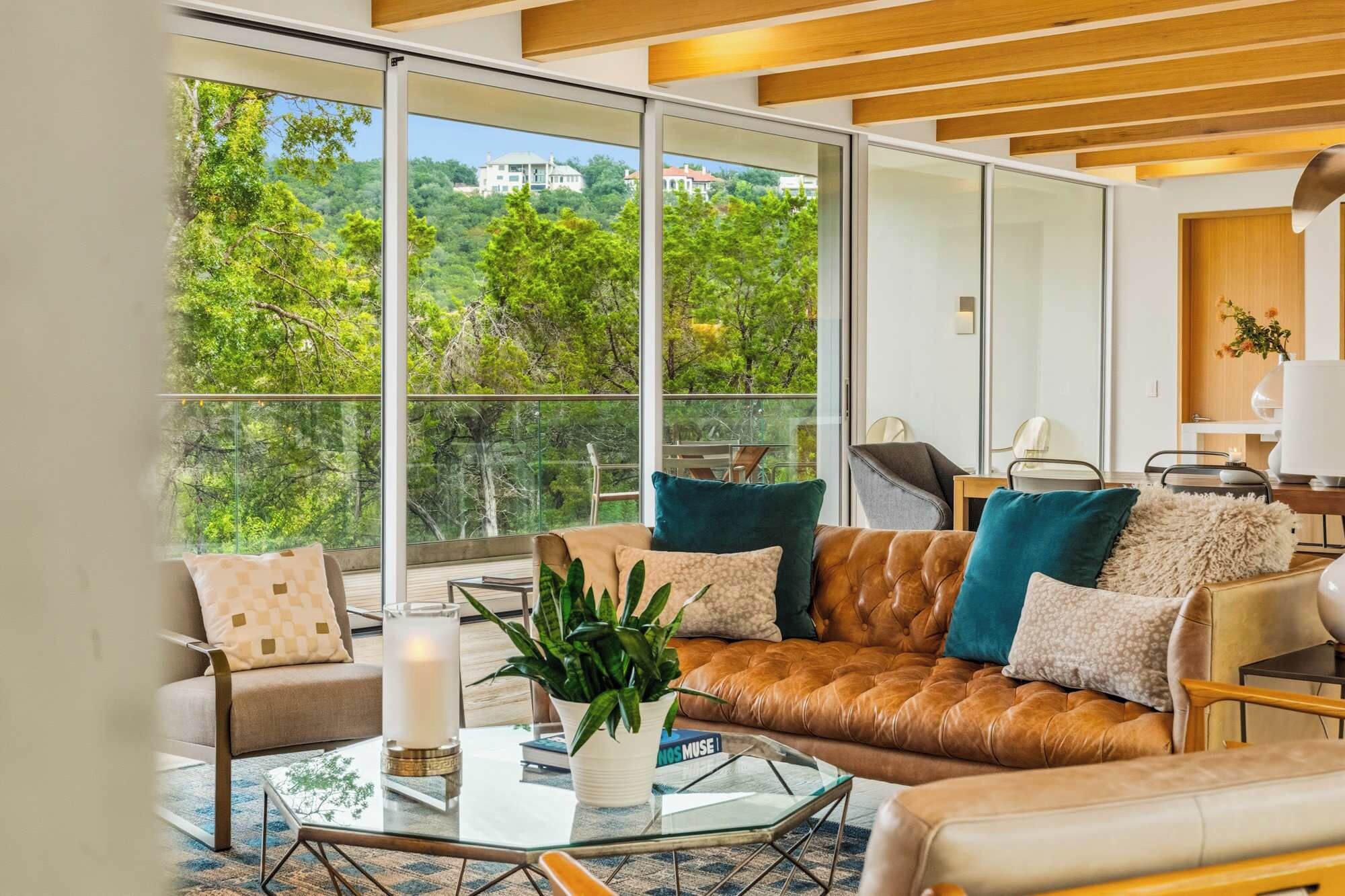 Un salon moderne avec un canapé en cuir, une table basse en verre et une vue sur un paysage verdoyant grâce à de grandes fenêtres.