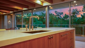 A modern kitchen with a large island, wooden cabinets, and a view of trees through large windows.