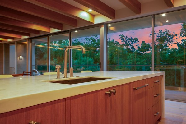 A modern kitchen with a large island, wooden cabinets, and a view of trees through large windows. - Thanksgiving in Austin | Chef’s Kitchen + Hot Tub (West Lake Hills)