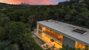 A modern house with a flat roof, large windows, and a view of a city skyline at sunset.