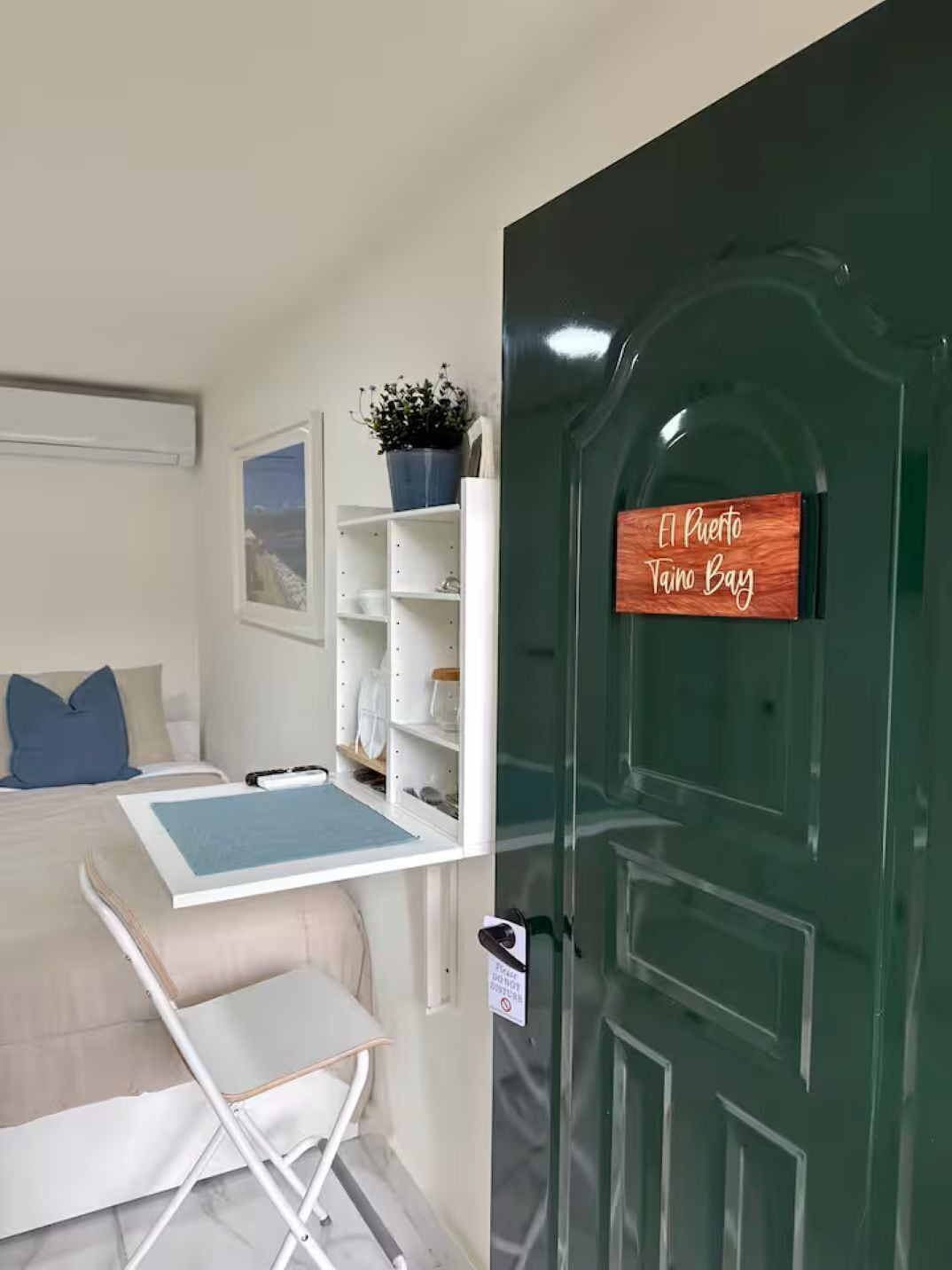 Une chambre d’hôtel comprenant un lit, un bureau, une chaise, une petite table, une étagère avec des objets décoratifs, une porte avec une pancarte et une fenêtre donnant sur un paysage.