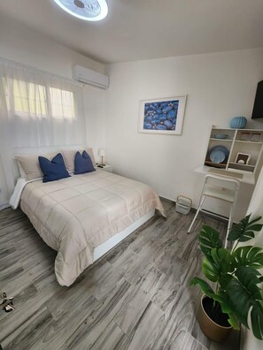 A bedroom with a bed, a bedside table, a wall-mounted air conditioner, a framed artwork, a shelf with decorative items, and a potted plant.