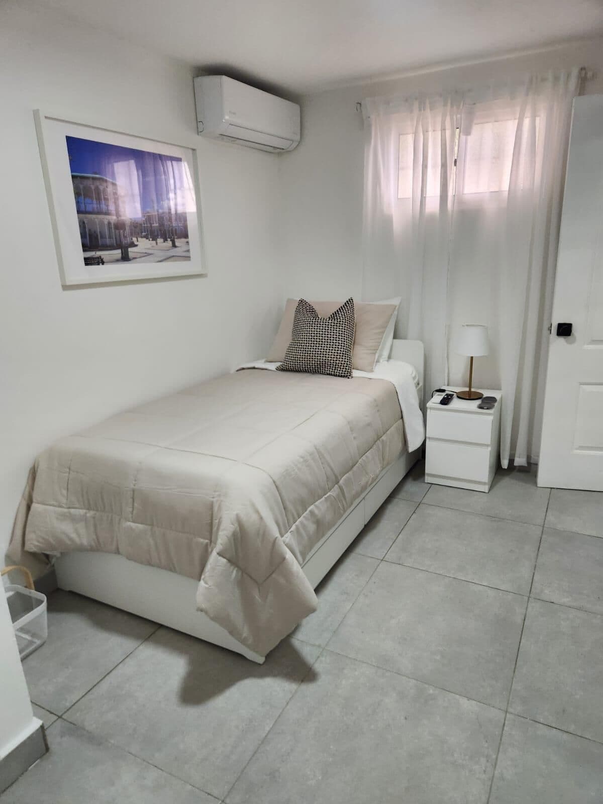 Un dormitorio con cama, mesita de noche, lámpara, aire acondicionado y un cuadro enmarcado en la pared.