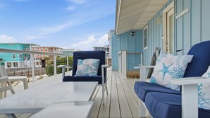 Un espace extérieur abrité, aménagé avec un canapé et des fauteuils bleus, une table, et offrant une vue sur la plage et les bâtiments.