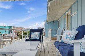 A covered outdoor seating area with a blue sofa and chairs, a table, and a view of the beach and buildings. - Steps to the Beach • Gulf + Sound Views (Navarre Beach)