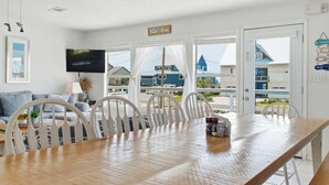 Un espace repas comprenant une table en bois, des chaises blanches, un téléviseur et une vue sur les bâtiments environnants.