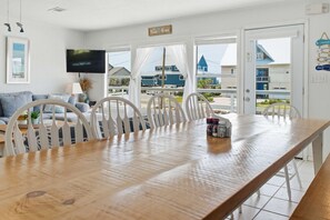 A dining area with a wooden table, white chairs, a television, and a view of buildings outside. - Steps to the Beach • Gulf + Sound Views (Navarre Beach)