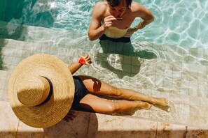 Pessoa de biquíni e chapéu de palha à beira de uma piscina com água cristalina.