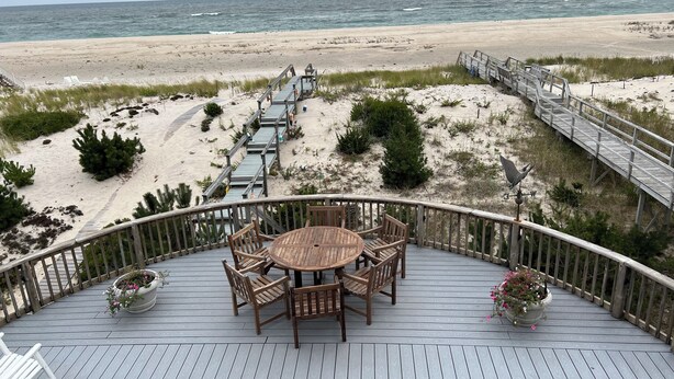 Eine Holzterrasse mit rundem Tisch und Stühlen mit Blick auf einen Strand und eine Holzpromenade.