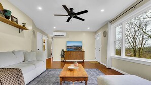 A living room with a sofa, a wooden table, a ceiling fan, and a large window with a view. - Hickory Ridge Homestead | Free Farm Tours + Firepit (Augusta)