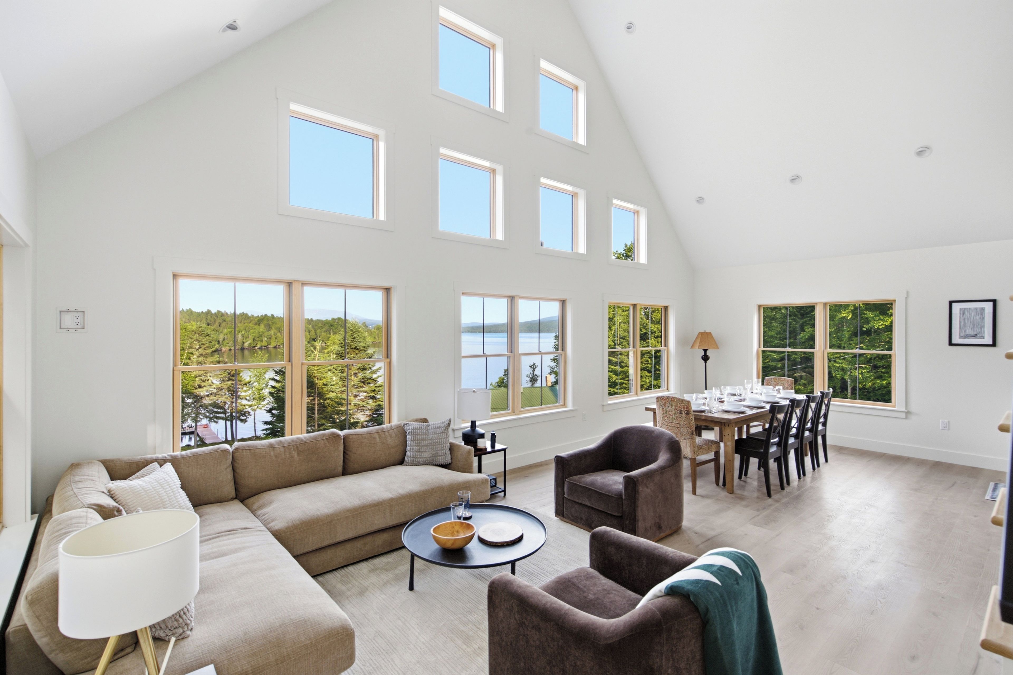 A spacious living room with a high ceiling, large windows, a sofa set, a dining table with chairs, and a view of a lake.