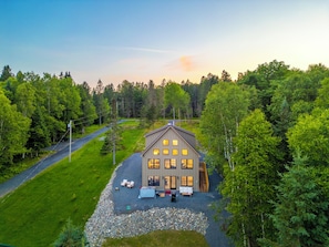 A two-story house with a driveway leading to a front porch, surrounded by a forested area. - Lakefront Modern Farmhouse with Private Dock (Rangeley)