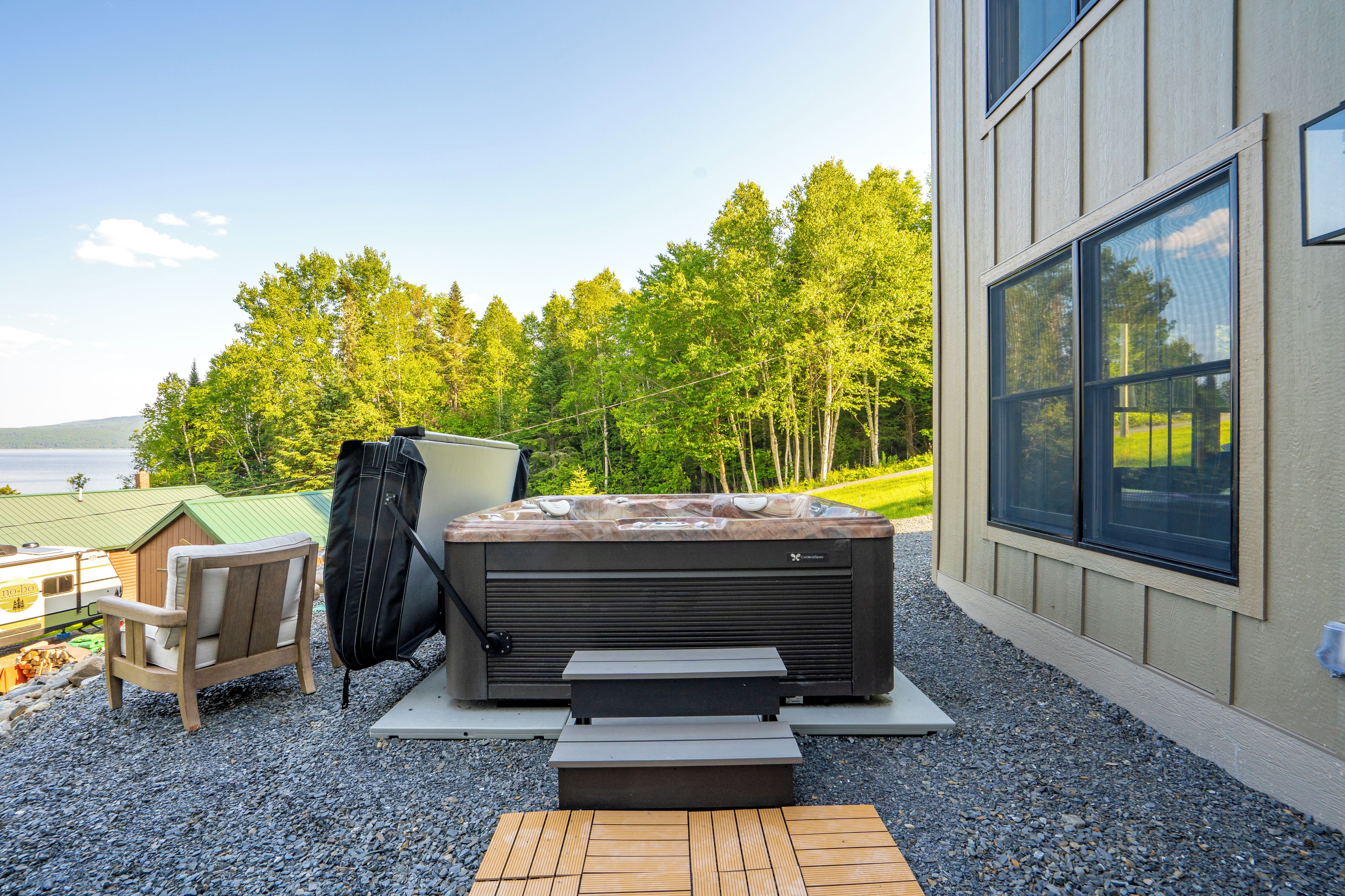 Un bain à remous avec des marches, une chaise et une table, le tout installé sur une surface de gravier, avec vue sur des arbres et un bâtiment en arrière-plan.