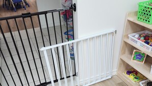 A room with a white baby gate, a wooden floor, a small table with chairs, and a shelf with toys.