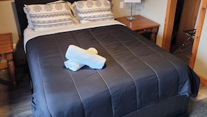 A bedroom with a wooden ceiling, a bed with a black mattress and patterned pillows, a bedside table with a lamp, and a wooden floor.
