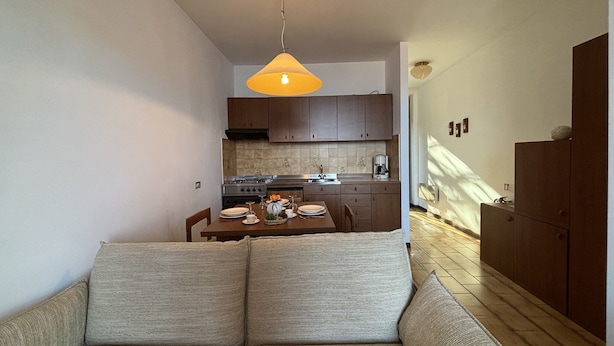 Un espace de vie compact comprenant un canapé, une table à manger pour deux personnes, un coin cuisine avec des placards et des appareils électroménagers, et un luminaire suspendu.