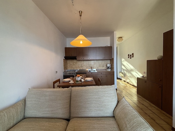 Ein kompakter Wohnraum mit Sofa, Esstisch für zwei Personen, Küchenzeile mit Schränken und Geräte sowie einer Pendelleuchte.