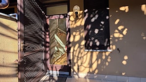 Une grille métallique fermée, une porte en bois, une petite fenêtre avec des volets et un sol carrelé.