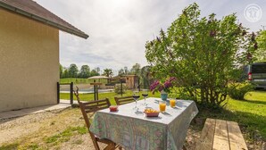 En udendørs spiseplads med et bord dækket til to, omgivet af grøn vegetation og en bygning.