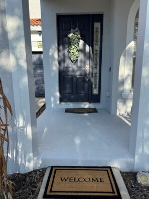 Una puerta principal con un felpudo que dice 'WELCOME', un timbre y un adorno de guirnalda.