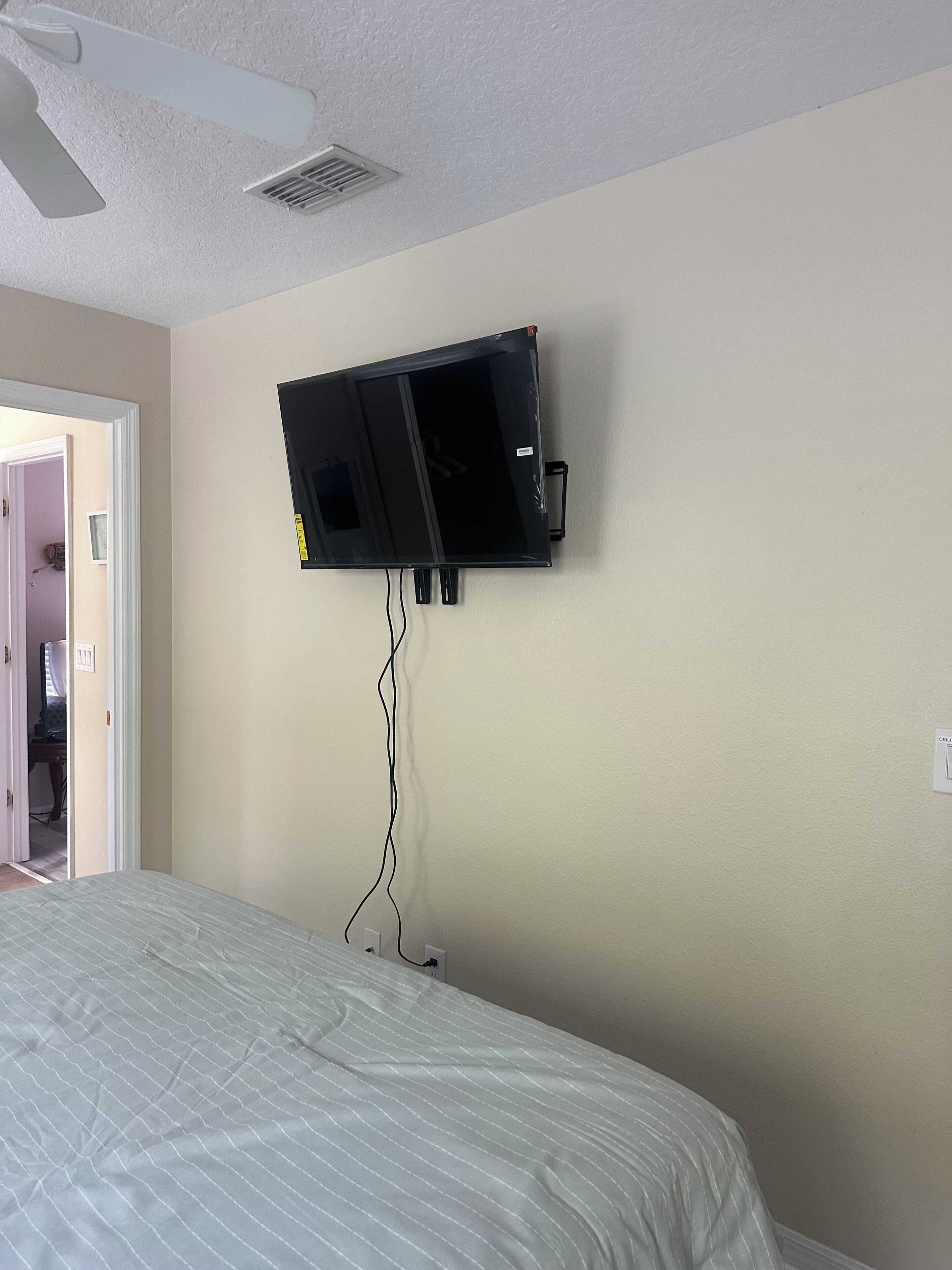 Ein Schlafzimmer mit einem Bett, einem an der Wand befestigten Fernseher, einem Deckenventilator und einer Tür, die in einen anderen Raum führt.
