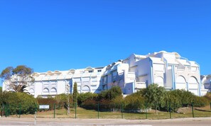 Ein großes, weißes Gebäude mit mehreren Balkonen und Bogenfenstern, umgeben von viel Grün und blauem Himmel.