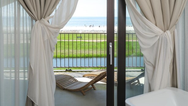 A balcony with a view of a beach, a wooden lounge chair, and a white bathtub. - Water's Edge Wellness Retreat With Sauna (Workum)