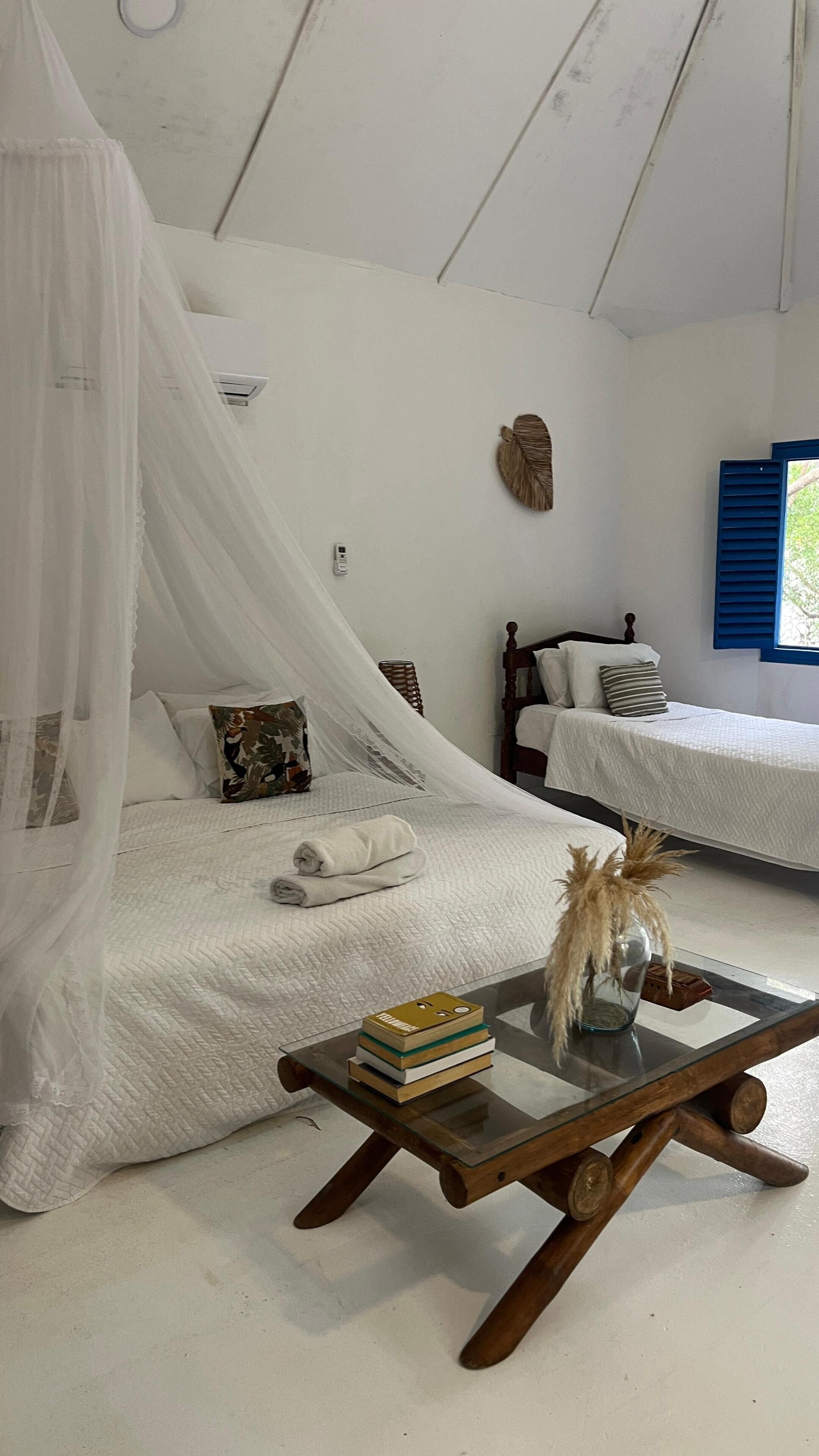 A bedroom with a canopy bed, a bedside table with books, a wall-mounted heater, and a window with blue shutters.