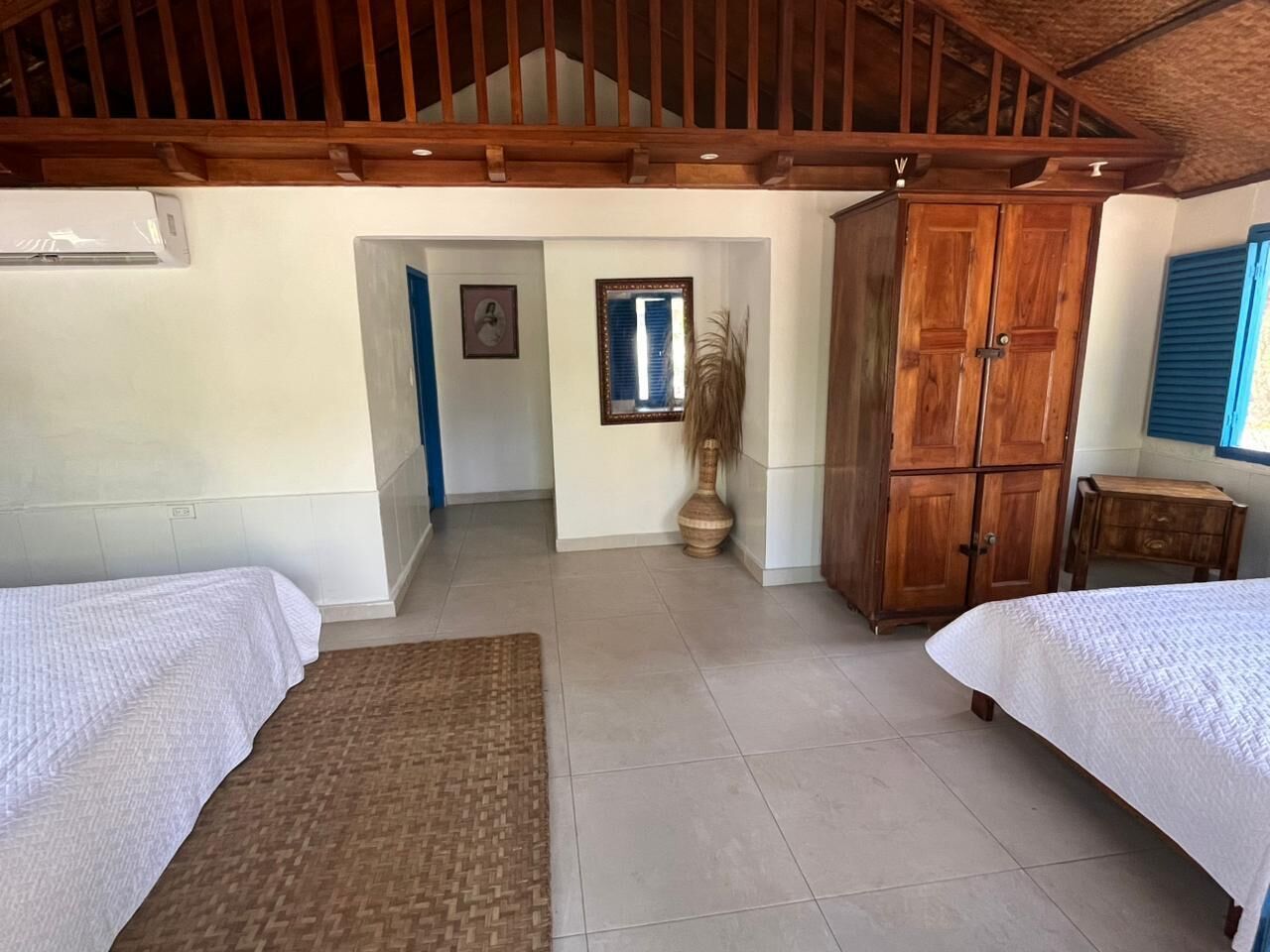 A bedroom with two beds, a wooden wardrobe, a large potted plant, and a staircase leading to an upper level.