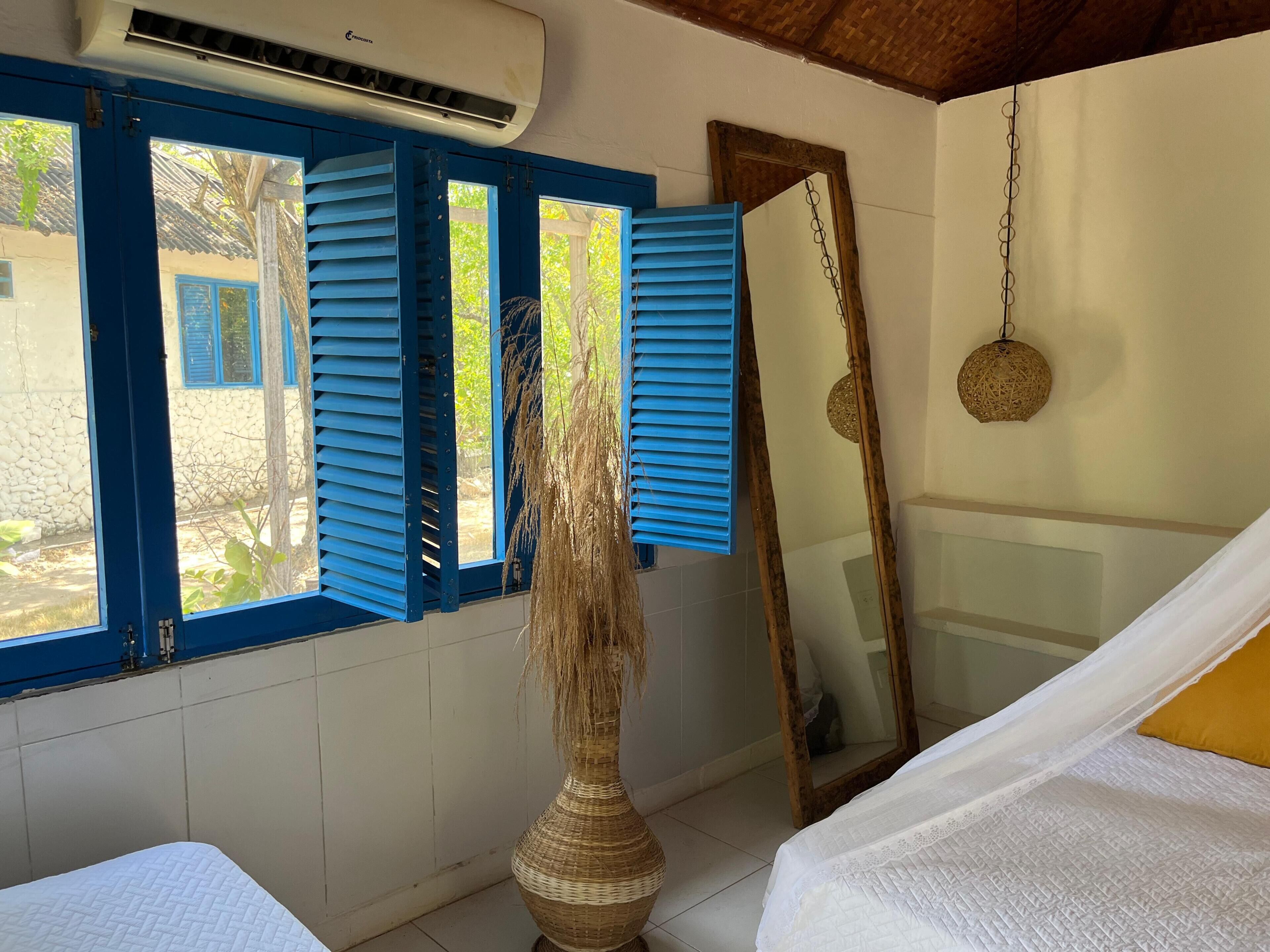 Una habitación con una cama, un espejo grande, un jarrón con plantas secas y una ventana con contraventanas azules.