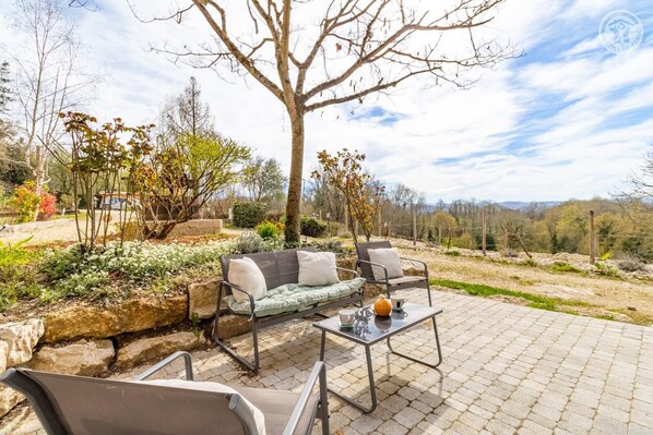 Une terrasse extérieure aménagée avec un canapé, des fauteuils et une table, entourée de plantes et offrant une vue sur la campagne.