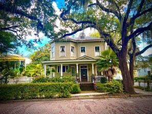 A two-story house with a porch, surrounded by lush greenery and a large tree.