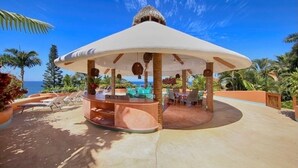 A covered outdoor seating area with a thatched roof, surrounded by palm trees and a view of the ocean. - Private Sayulita Estate 3 Villas, 3 Pools, Ocean Views, Walk to Beach, Sleeps 20 (Sayulita)