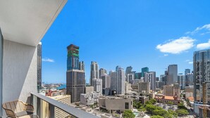 Paisagem urbana com edifícios altos, céu claro e uma varanda com cadeira de vime.