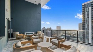 Un balcon avec des sièges d’extérieur, une vue sur la ville et, au fond, un bâtiment sur lequel on peut lire l’inscription « À LOUER ».
