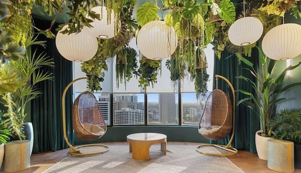 A room with two hanging chairs, a round table, and a city view through large windows. - 501 First #2109|Miami City Downtown Studio (Miami)
