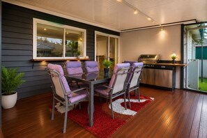 An outdoor dining area with a glass table and chairs, a grill station, and a view of the backyard.