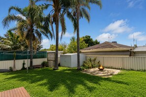 A backyard with a fire pit, palm trees, and a fenced area with a house in the background. - Cosy White Hills Luxury very close to Bendigo (White Hills)