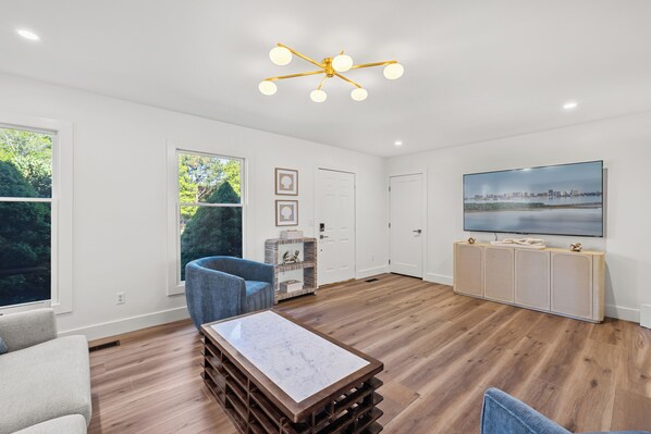 A spacious living room with a large TV, wooden flooring, a blue chair, and a marble coffee table. - Coastal Retreat in Peaceful Waterfront Community (Harbeson)
